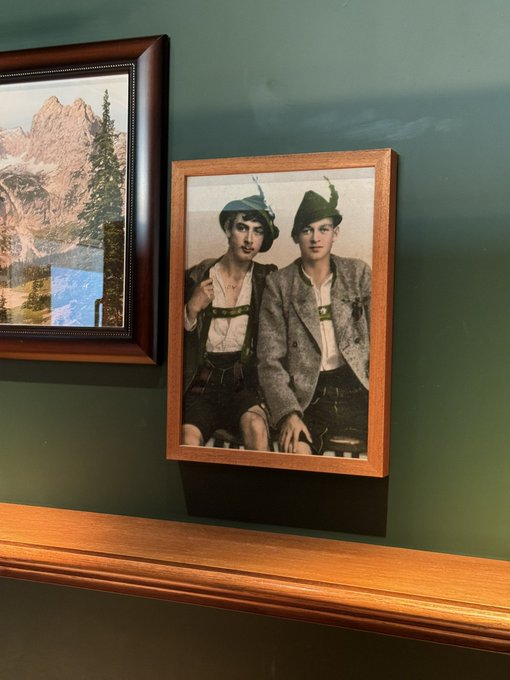 A framed vintage black-and-white photograph of two young men in traditional Bavarian or Alpine attire, displayed on a dark green wall next to a landscape painting.