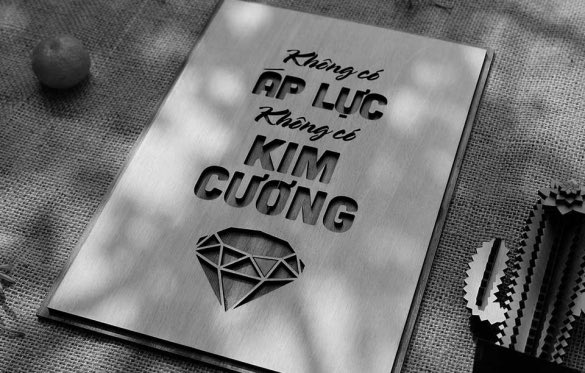 A grayscale photograph of a book or print titled 'Không có Áp Lực Không có Kim Cương' (No pressure no diamond) featuring a diamond graphic, resting on a textured surface.