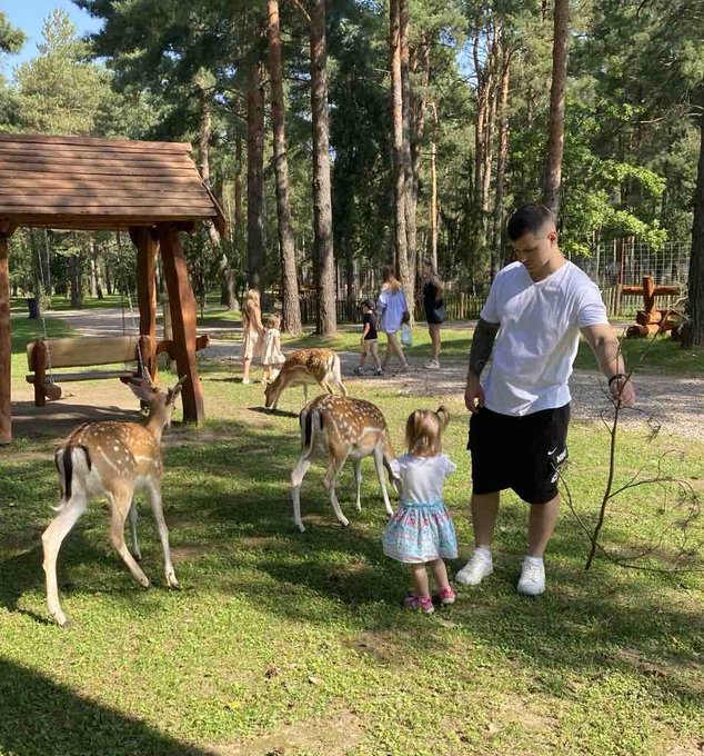 Family photo in a park or animal sanctuary where a man in a white shirt and black shorts stands with a young girl feeding fallow deer, with a wooden structure and trees in the background.