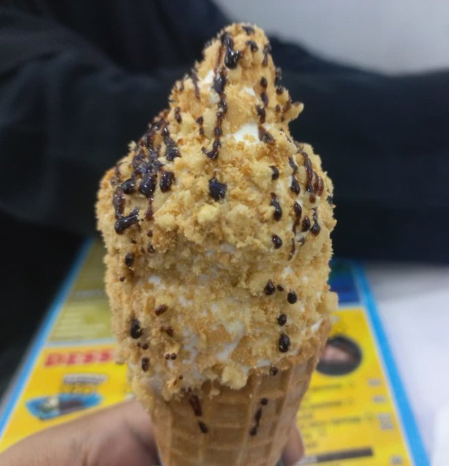 A hand holding a soft-serve ice cream cone in a waffle cone, covered in crushed peanut or cookie crumbs and drizzled with chocolate sauce.