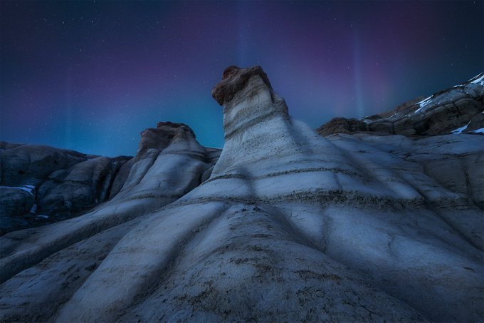A night landscape photograph of eroded rock formations under a dark blue sky illuminated by the Northern Lights and stars.
