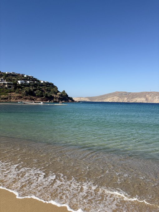 A serene beach scene with crystal-clear turquoise water gently lapping onto golden sand, framed by a hillside dotted with white Mediterranean-style buildings under a cloudless blue sky. The tranquil seaside view perfectly complements the post's celebratory mood, providing a luxurious backdrop to mark the SLX airdrop's growth to $32,565.