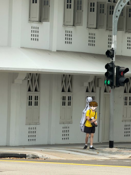 A street art mural of an anime-style character in a yellow shirt carrying a cello case stands at a pedestrian crossing next to a traffic light against a pale green building.