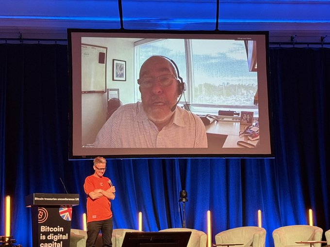 The image shows a man standing behind a podium, giving a presentation or speech at an event. He appears to be wearing headphones, suggesting that he might be using a microphone for his talk. The setting includes a large screen displaying a video of another person's face and body, which is likely part of the presentation. 

The image also contains text that reads "Bitcoin To 140k by the end of the year". This could possibly be related to Bitcoin or cryptocurrency, as it seems to be a topic of discussion during the event. The presence of chairs in the background indicates that there might be an audience present for this presentation.