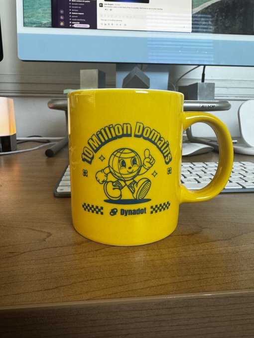 A bright yellow 'Dynadot' branded coffee mug featuring a cartoon astronaut mascot and the text 'Competition Domination' displayed on a wooden desk.