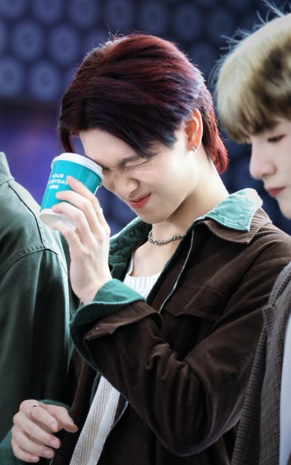 A young man with reddish-brown hair and a brown jacket with green collar smiles while holding a turquoise paper cup to his face, with another person partially visible beside him.