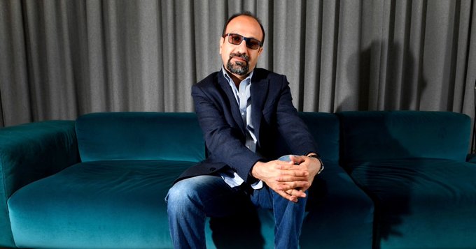 A man with a beard, wearing sunglasses, a dark blazer, and jeans, sits with his hands clasped on a teal velvet couch against a backdrop of gray curtains. The image appears to be a portrait of Asghar Farhadi, the Iranian filmmaker referenced in the accompanying post, which criticizes his silence regarding the 2017-18 Iranian protests and his acknowledgment of the Oscar award's connection to Iranian people.
