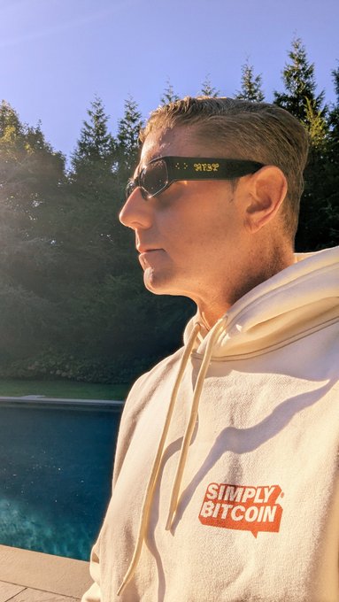 A side profile photo of a man with short hair wearing black sunglasses displaying a Bitcoin ticker price and a cream hoodie with a red 'SIMPLY BITCOIN' logo, taken outdoors near a pool.