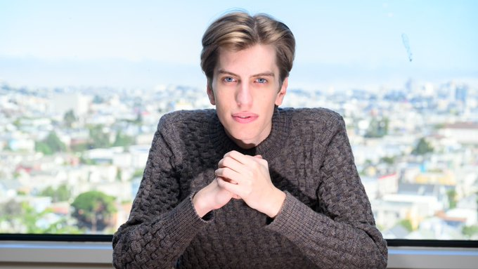 A young man with light brown hair wearing a gray knit sweater, posed with his hands clasped against a backdrop of a city skyline.