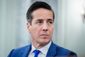 A man in a blue suit and tie looks off-camera with a serious expression, appearing to listen intently during what seems to be a formal proceeding or interview related to the CLARITY Act discussion.