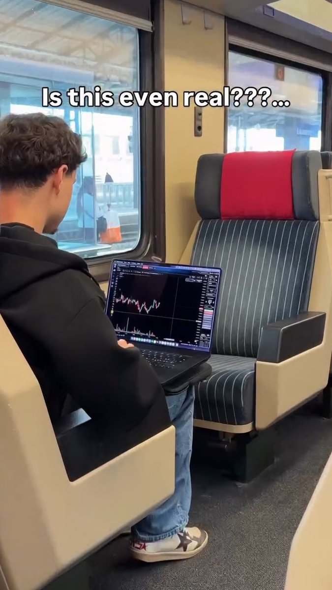 A man sits on a train with a laptop displaying a stock or crypto trading chart, with overlaid text asking "Is this even real???..."