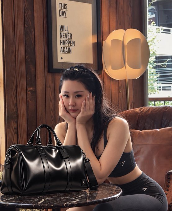 A young woman poses with her face resting in her hands next to a black leather duffle bag, with a framed poster reading 'THIS DAY WILL NEVER HAPPEN AGAIN' behind her.