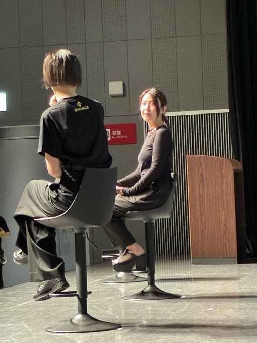 A woman smiling warmly while seated next to someone wearing a Binance t-shirt, backstage at an event, related to the "#JBWS2026" conference featuring tagged guests.