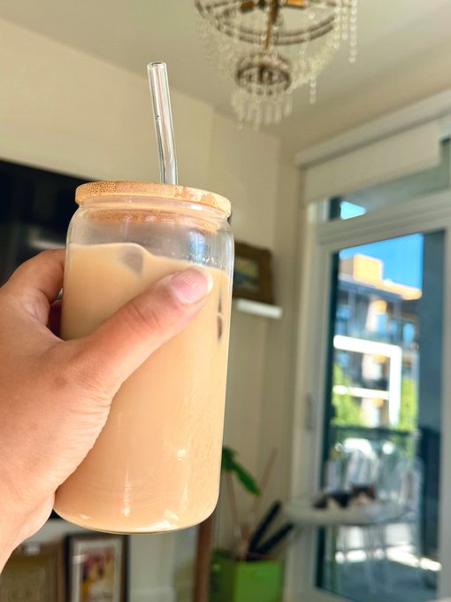 A hand holds up a glass jar filled with a light brown iced coffee drink topped with foam, complete with a glass straw and wooden lid, set against a sunny home interior with a chandelier.