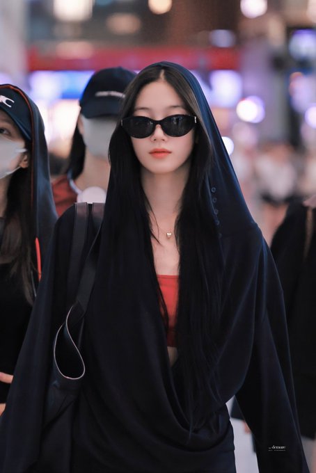 A young Asian woman wearing dark sunglasses and a black draped hooded outfit over a red top, posing in a crowded indoor venue.