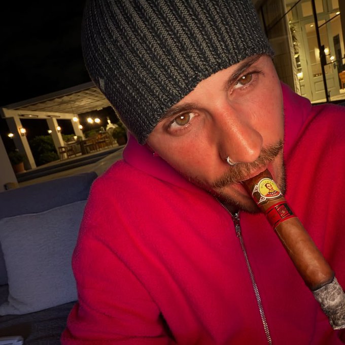 A man wearing a gray beanie and red zip-up jacket with a nose ring smoking a cigar in an outdoor evening setting.
