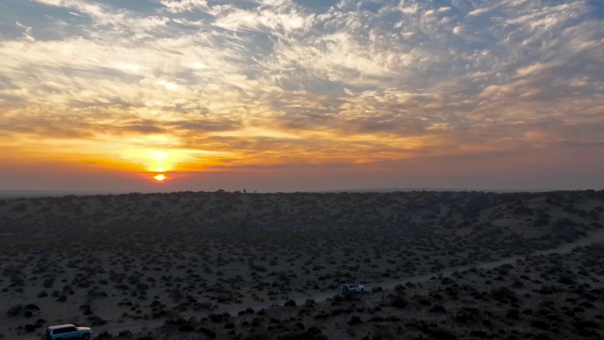 The image features a beautiful sunset over a desert landscape with a few clouds scattered across the sky. In the foreground of the photo, there are two cars parked near each other, possibly enjoying the view or heading home after a day's work. The scene is captured from an aerial perspective, providing a unique and captivating view of the desert and its surroundings.