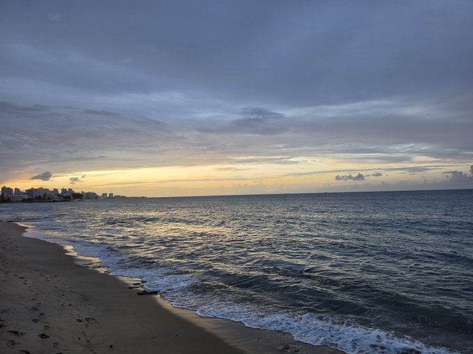 A serene sunset view over the ocean, with gentle waves lapping against a sandy beach and a distant city skyline silhouetted on the left horizon, perfectly capturing the tranquil "Great night here" vibe.
