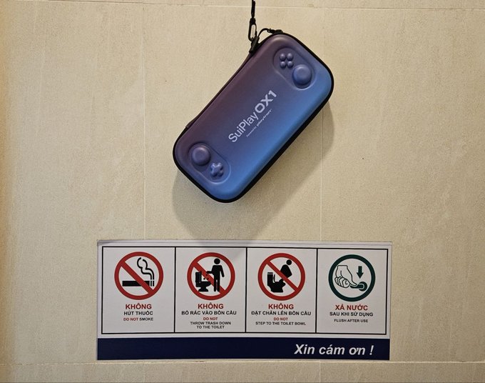 A 'SulPlay OX1' portable gaming device hanging on a tiled wall above a sign with bathroom instructions in Vietnamese ('KHÔNG HÚT THUỐC', etc.) and 'Xin cảm ơn!'.