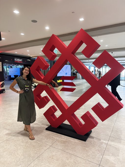 A woman in an olive-green dress poses next to a large red geometric knot sculpture in a modern indoor venue with a Summit sign visible in the background.