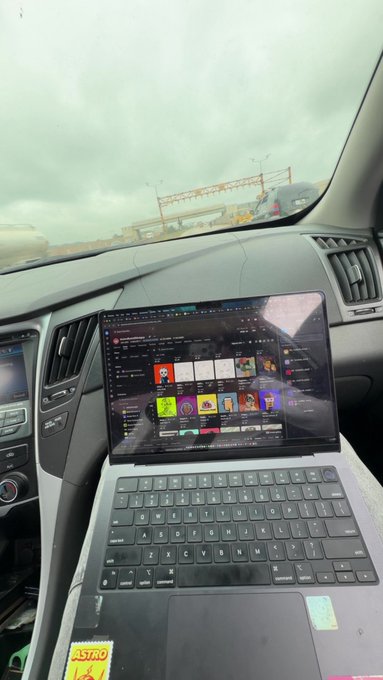 A laptop placed on the passenger seat of a car displaying a website with various items/images, with a view of a construction site and cloudy sky through the windshield.