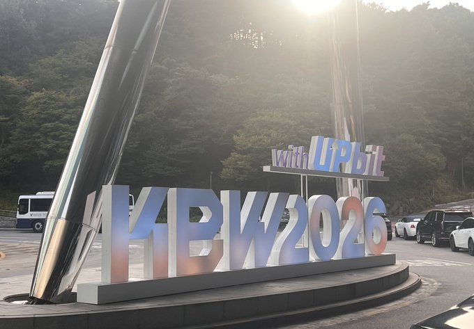 A large outdoor installation displays the text "with UPbit" and "KBW2026" in 3D letters, set against a mountainous backdrop with vehicles parked nearby.
