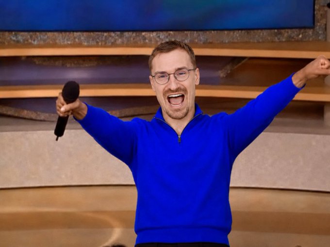 A man wearing glasses and a blue sweater smiles joyfully with arms raised, holding a microphone against a neutral background.
