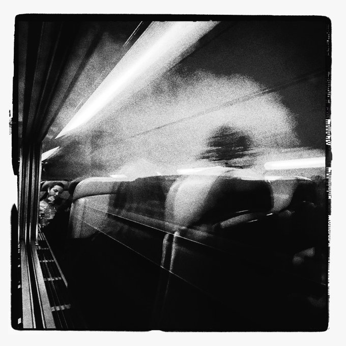 A grainy black-and-white photograph taken inside a subway car, showing a blurred figure seated along the wall with bright streaks of light reflecting off the windows.