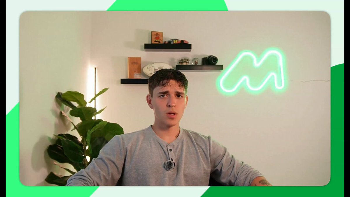 A man wearing a grey sweatshirt with neon green lettering on his shirt is sitting down. There's an unusual neon sign hanging above him, which reads "M". The background appears to be a room with several objects such as potted plants that are arranged in different areas of space within the scene. On top of one of these plants, there seems to be a clock or other decorative item. The man is looking at the camera, and this image has text overlaying it that says "Global remittances still cost ~6.36%. Move money on Morph for a fraction of that."