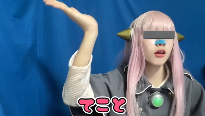 A person wearing a pink wig and gray outfit poses with their arm raised against a blue background, with pink Japanese text at the bottom.