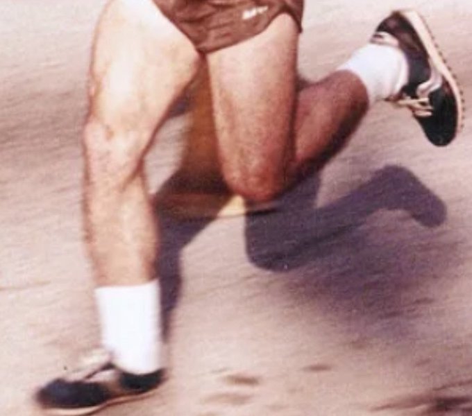 Close-up of a runner's legs in mid-stride wearing white socks and dark running shoes, with the other leg appearing to bend unnaturally behind, suggesting an injury or visual illusion.