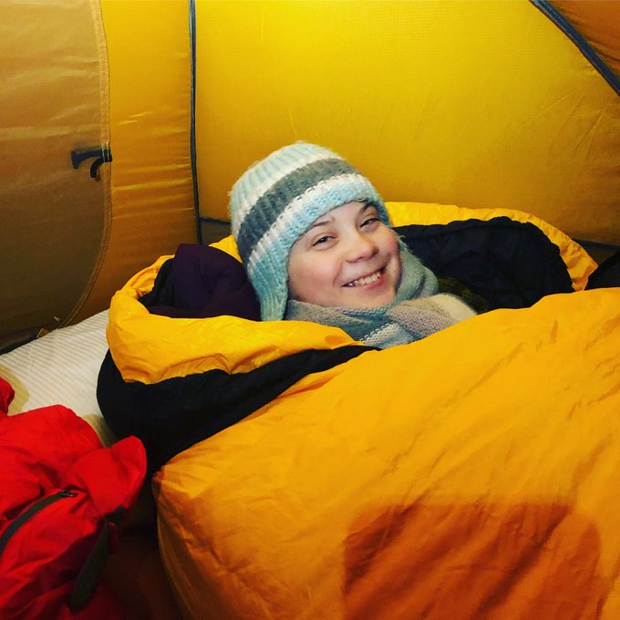 A smiling young person wearing a striped knit hat and scarf, wrapped in a yellow sleeping bag inside a yellow tent.