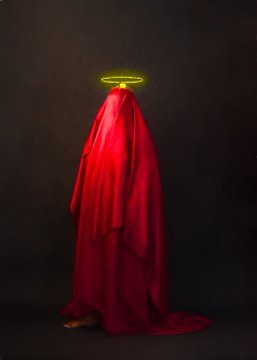 The image features a person standing against a black background, wearing a red cape or dress that covers their entire body except for their head. The person's face is not visible due to the covering of the cape. The caption reads "Saint Without a Name | To Hold Light in Shadow". This phrase suggests that the person might be associated with a saintly figure who is known for holding onto light and shadows, symbolizing hope or guidance during difficult times.