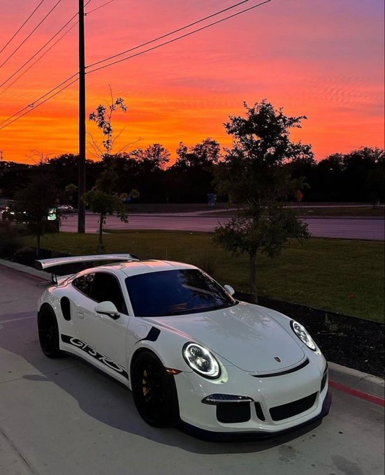 A white Porsche 911 GT3 RS is parked outdoors against a vibrant orange and pink sunset sky.