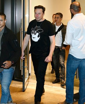 A man, believed to be Elon Musk, walks through a doorway wearing a black t-shirt featuring the Dogecoin Shiba Inu mascot wearing a space helmet. He is surrounded by several other individuals in what appears to be a public or professional setting.