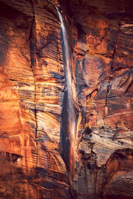 A natural rock formation featuring a narrow slot canyon with layered, textured walls in shades of orange, brown, and gray, illuminated by sunlight filtering down from above.