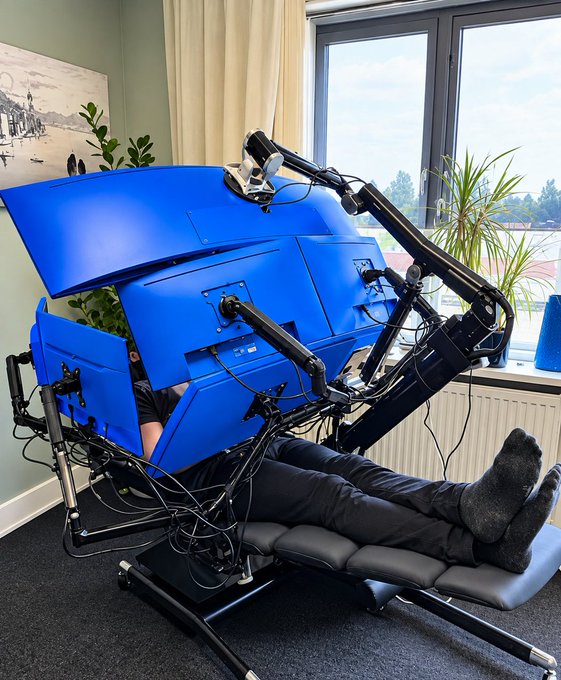 A man reclines on a black lounge chair wearing a large, blue, cube-shaped helmet equipped with multiple camera arms and cables, seemingly for immersive monitoring. The image humorously suggests he is using this elaborate, high-tech rig just to obsess over his modest $67 crypto portfolio, staying intensely "locked in" on the markets.