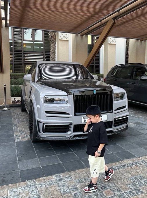 A young boy wearing sunglasses, a black shirt, and sneakers stands in front of a two-tone (white and black) Rolls-Royce SUV.