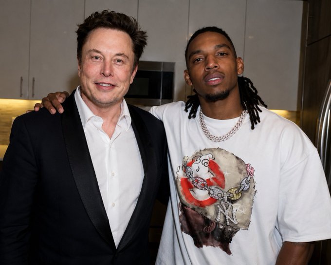 The image shows two men standing next to each other, with one man wearing a suit and the other wearing a t-shirt featuring a graphic of a face. The man in the suit has his arm around the other man's shoulder, while they both pose for the camera. In the background, there is a refrigerator visible on the right side of the image.

The caption to the image reads: "Sources confirm B Lou was the witness who signed Elon Musk’s paperwork to become 'Dogeus Maximus' @BLouu Is this shit true?". This statement suggests that someone else might have signed the papers for the two men, and it is likely related to a famous person or event.