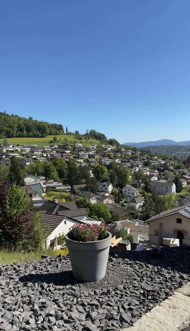 A scenic photograph of a town in a green valley viewed from a hilltop, featuring a foreground of gray gravel and a pot of pink flowers under a clear blue sky.
