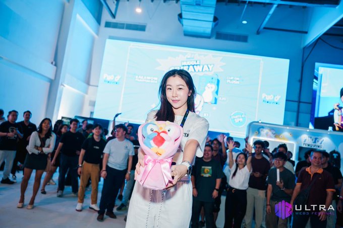 Event photograph of a young woman in a white outfit holding a pink heart plush toy with an Angry Birds face, standing in front of a crowd and a large screen at a gaming or anime convention, watermarked 'ULTRA ESPORTS'.