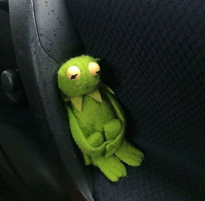 A green Kermit the Frog plush doll with glowing eyes sitting on a dark fabric car seat.