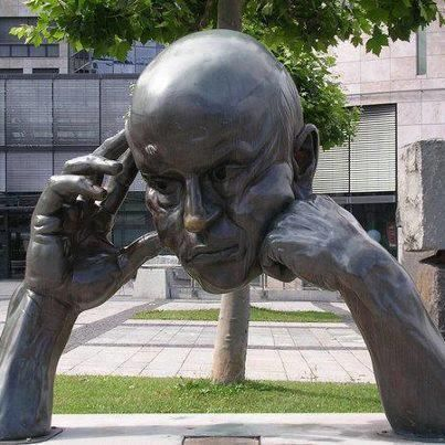 urn-shaped sculpture of a bald man's head, with hands on chin and eyes closed, stands against a backdrop of trees and buildings.