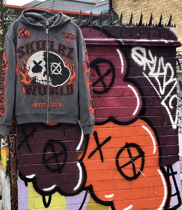 A grey zip-up hoodie featuring skull graphics and 'SKULLZ WORLD EST. 2022' text with flame designs, displayed against a graffiti-covered brick wall.