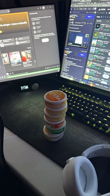A stack of round containers labeled 'TROPICAL ICE' and 'SHERB' sits on a desk in front of two computer monitors, with white headphones resting nearby.