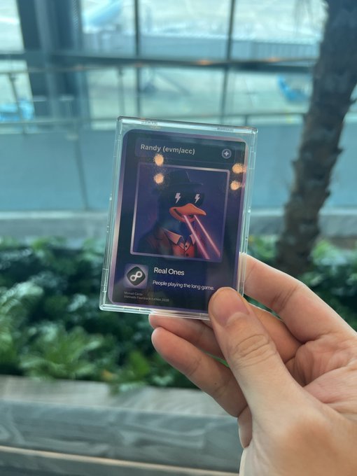 A hand holds up a plastic-encased "Real Ones" card featuring the wallet address "Randy (evm/acc)" with the tagline "People playing the long game," earned as a keepsake from a Monad meetup.