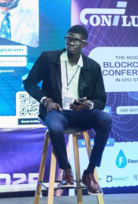 A man wearing glasses, a black jacket, white polo shirt, and lanyard sits on a wooden stool at the 'CONFLU BLOCKCHAIN CONFERENCE IN OYO STATE,' holding a phone, with sponsor logos like Quorum and and a QR code visible in the background.