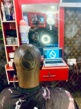 A man with a freshly shaved head sitting in a barbershop, facing a mirror with a ring light, with a laptop displaying a water drop logo on the counter.
