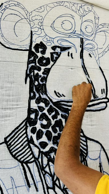 A close-up of an artist's arm working on a textile piece, sketching and painting the outlines of a giraffe with black and blue markers on a gridded fabric.