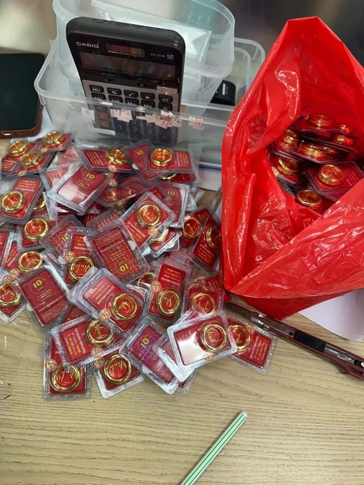 A pile of gold coin-shaped items in red and clear packaging on a desk, spilling out of a red plastic bag, with a Casio calculator visible in the background.
